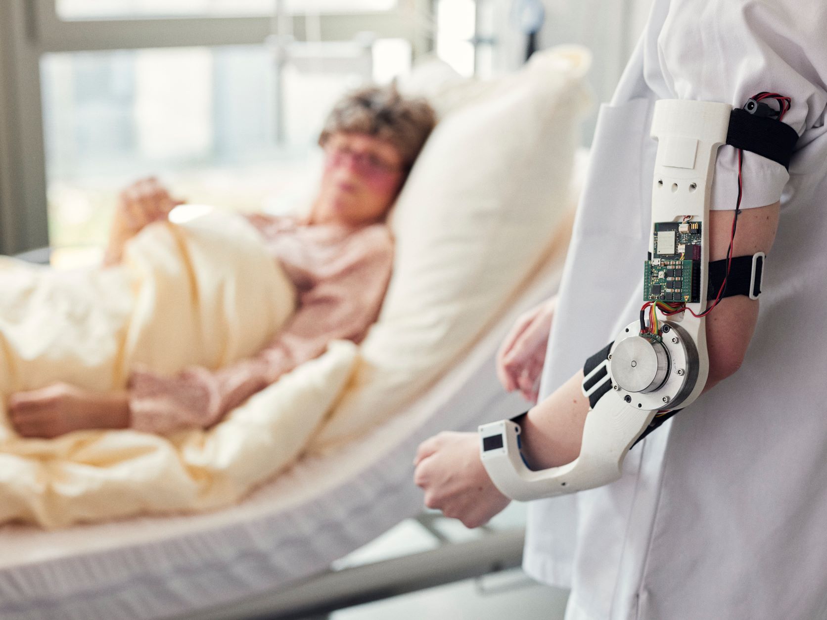 Eine Person trägt ein technisches Gerät am Arm. Im Hintergrund liegt eine ältere Frau im Bett.